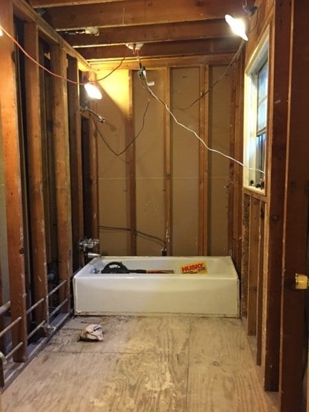 Bathroom demolished to the studs with the bathtub still in place and work lights hung from the ceiling joists