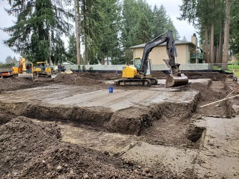 Excavator beside a residential foundation pad dug out to grade, with the spoil benched around the edge