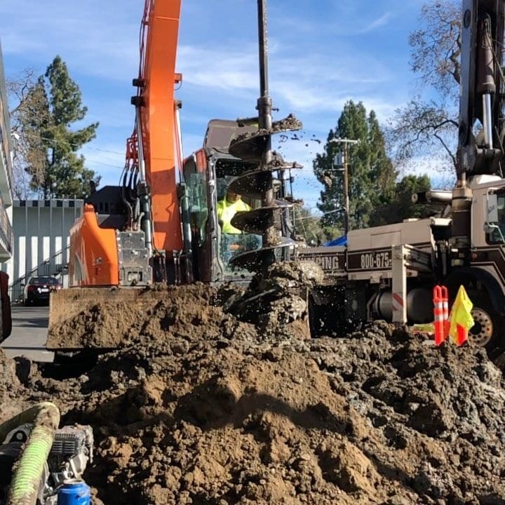 Large auger on an excavator drilling a foundation pier, with spoil coming up the flights and a crew member in high-visibility clothing